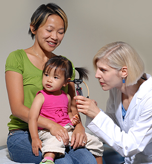 Toddler sitting on woman's lap while doctor examines the toddler's ear with otoscope.